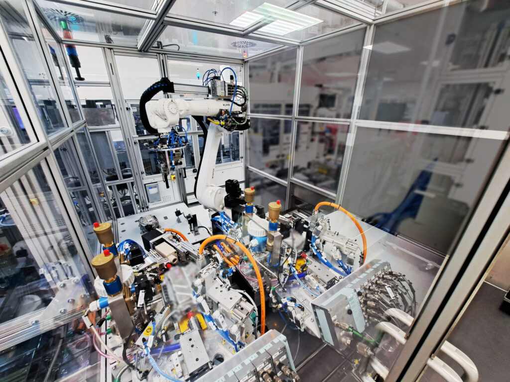 A robotic arm operates inside a glass-enclosed automated assembly station with various mechanical components and wiring.