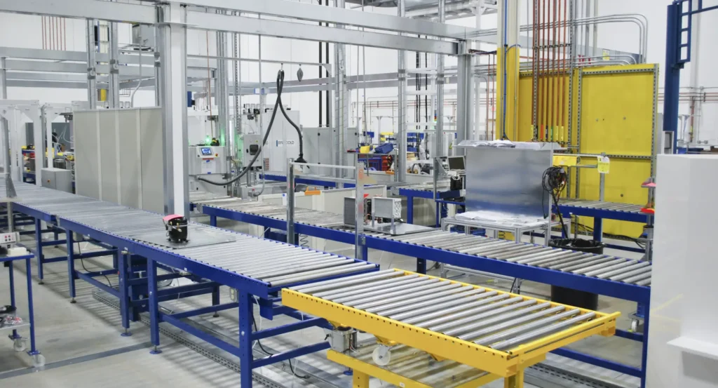 Automated conveyor system in a factory setting with metal rollers, machinery, and various equipment visible in a clean, industrial environment.
