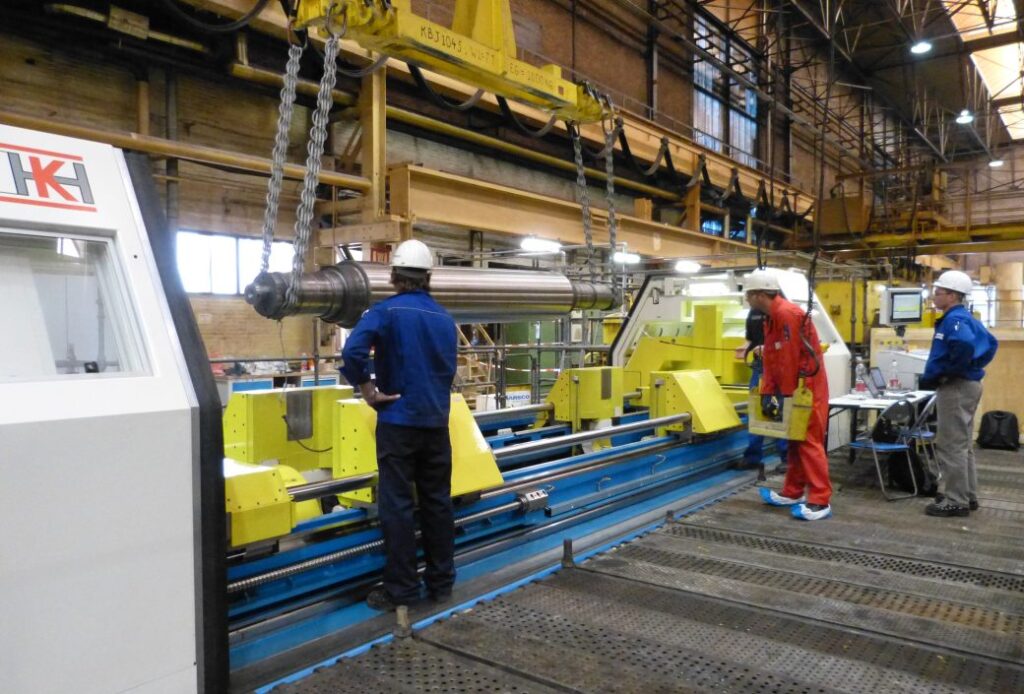 Three workers in safety helmets operate and inspect large industrial machinery inside a spacious factory or manufacturing facility.