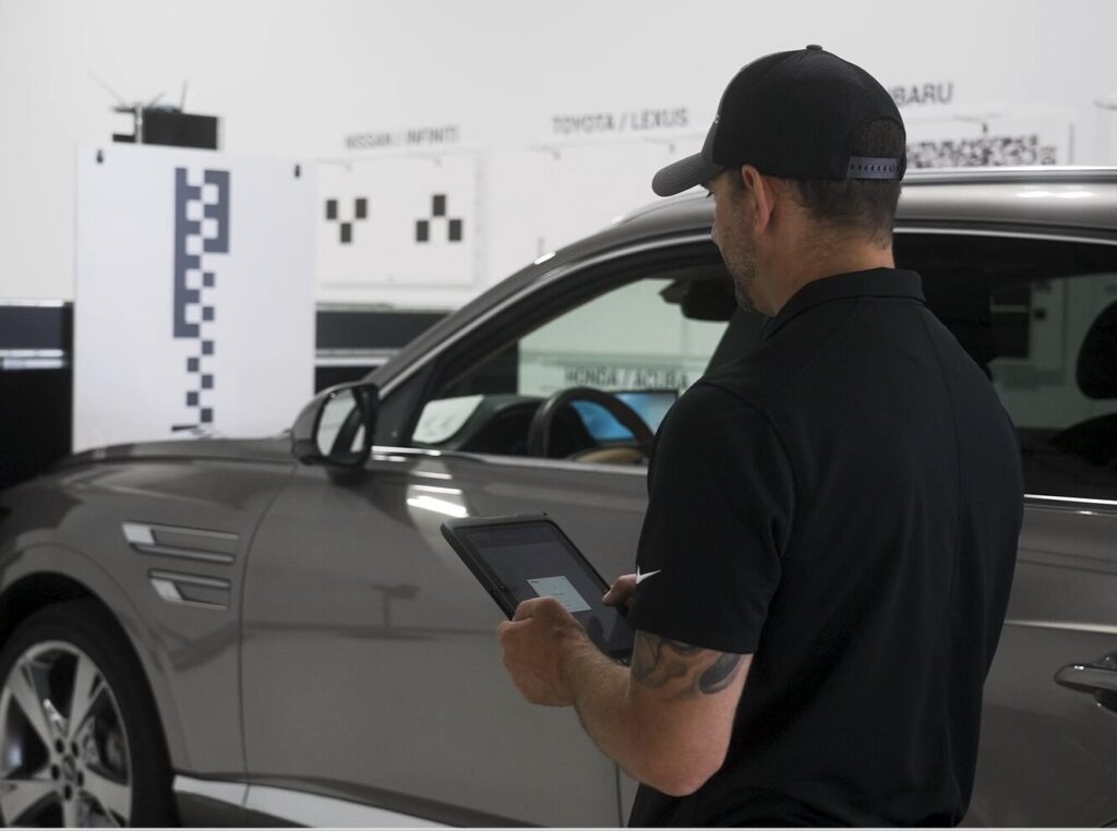 A man wearing a black shirt and cap uses a tablet while standing next to a gray SUV in an automotive workshop.